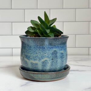 May include: A ceramic planter with a mottled blue and green glaze, featuring a flared rim and a matching circular saucer. A small green succulent plant with thick, pointed leaves is potted inside the container, which has a smooth finish.