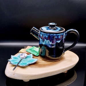 May include: A ceramic teapot with a blue and black glaze, a matching lid, and a curved handle. It sits on a wooden serving board with a leaf-shaped dish and a small spoon. Tea bags are also on the board.