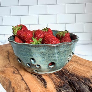 May include: A round, speckled teal ceramic colander bowl with a wavy rim and multiple drainage holes, filled with fresh red strawberries. The bowl features a rustic, hand-crafted appearance with visible variations in the blue-green glaze.