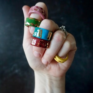 May include: A hand displays a collection of colorful ceramic rings. The rings are in shades of pink, green, turquoise, burgundy, and yellow, with gold accents. The rings are of various sizes and have a glossy finish.