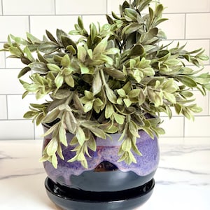Puede incluir: Una pequeña planta verde artificial con hojas puntiagudas en una maceta de cerámica redonda. La maceta tiene un acabado brillante con una sección superior de color morado intenso que se funde con una base negra sólida, sobre un plato negro a juego.
