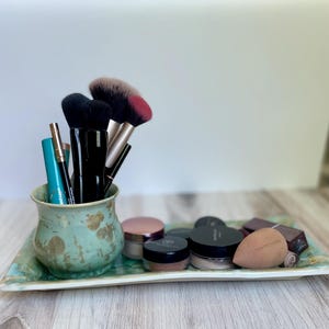 Puede incluir: Una colección de brochas de maquillaje y cosméticos dispuesta sobre una bandeja rectangular de cerámica vidriada turquesa. La bandeja contiene una variedad de productos de maquillaje, incluyendo tarros de polvos, una esponja de maquillaje y un pequeño recipiente de cerámica con brochas y aplicadores.
