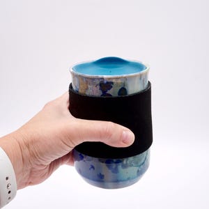 May include: A ceramic travel mug with a blue interior and lid, featuring a black sleeve. The mug has a unique, speckled glaze in shades of blue and green. The mug is designed for hot or cold beverages.