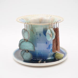 May include: A collection of handmade earrings displayed on a ceramic holder. The earrings feature various shapes and colors, including turquoise, copper, and green. The holder is a blue and white ceramic piece, with a matching saucer.