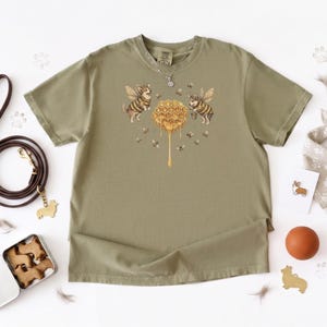 May include: An olive green short-sleeve t-shirt featuring a central graphic of two corgis dressed as bees flanking a dripping honeycomb, surrounded by small flying bees. The shirt has a crew neckline and a silver-toned chain necklace resting on it.