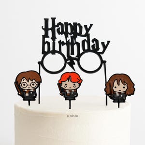Może przedstawiać: Czarny topper na tort z napisem "Happy Birthday" i okularami oraz trzema postaciami z kreskówek. Postacie mają brązowe, rude i brązowe włosy i trzymają różdżki. Topper na tort jest na białym torcie.