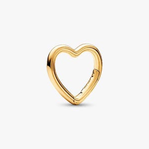 Puede incluir: Un charm en forma de corazón de tono dorado con un acabado liso y pulido. El charm cuenta con un mecanismo de apertura con bisagra funcional en el lado derecho, lo que permite sujetarlo a varios artículos. La superficie metálica refleja la luz de manera uniforme.