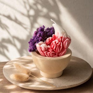 May include: A sculpted candle shaped like a blooming flower with white and red wax petals, featuring a single white wick. The candle is nestled in a small, beige, bowl-shaped container alongside a cluster of dried purple flowers and two tan stems.