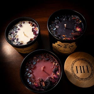 Puede incluir: Tres velas en recipientes negros con superficies de cera decoradas con pequeños cristales y pétalos de flores secas. Una vela es blanca, otra negra y otra roja oscura. La etiqueta dice in memoriam. Una tapa de madera presenta las letras HH.