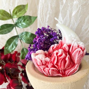 May include: A sculpted candle shaped like a blooming flower with white and red marbled wax, featuring a single white wick. The candle rests inside a beige, textured, bowl-shaped holder, accompanied by purple dried flowers and green leaves.