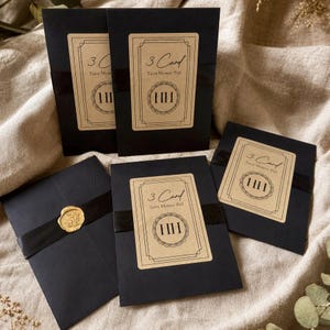 May include: Four black rectangular envelopes, each featuring a tan label with the text 3 Card Tarot Mystery Pull and the Roman numeral III inside a circular border. One envelope is sealed with a gold wax seal over a black ribbon band.