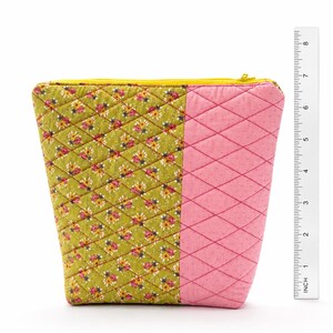 Könnte beinhalten: Ein gesteppter Beutel mit gelbem Reißverschluss. Der Beutel ist in zwei Bereiche unterteilt: einen mit einem grünen Blumenmuster und einen rosa mit einem Rautenmuster. Der Beutel ist etwa 10 cm hoch.