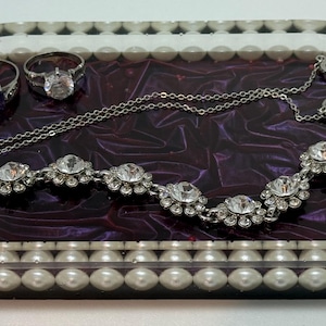 May include: An oval jewelry tray with a pearl border, holding a silver necklace, bracelet, and two rings. The tray's interior has a textured purple design, creating a luxurious display for the accessories.