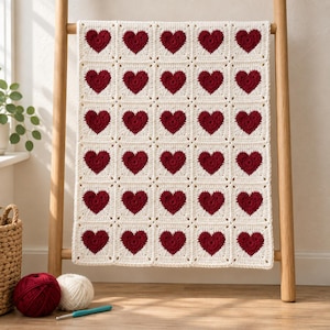 Peut inclure: Une couverture en crochet faite à la main composée d'une grille de trente motifs carrés, chacun présentant un cœur rouge foncé centré sur un fond crème. La couverture est posée sur une échelle en bois, avec des pelotes de laine rouge et blanche.