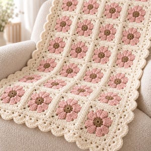 Peut inclure: Une couverture crochetée à la main présentant un motif répétitif de carrés. Chaque carré contient une fleur rose avec un centre marron sur un fond crème. La couverture est finie avec une bordure festonnée de couleur crème sur les bords.