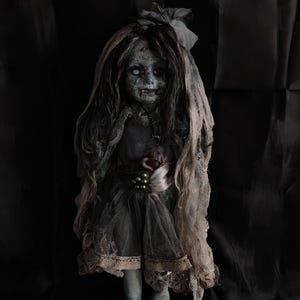 Puede incluir: Una muñeca antigua y espeluznante con tez pálida, ojos blancos y cabello oscuro. La muñeca viste un vestido gris andrajoso con encaje y un velo transparente. Una cinta con cuentas doradas adorna la cintura. La estética general es gótica y misteriosa.