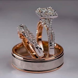 May include: A set of three rings, including a wedding band and two engagement rings. The engagement rings feature a square-cut diamond surrounded by smaller stones. The wedding band is two-toned, with a silver centre and a gold rim.