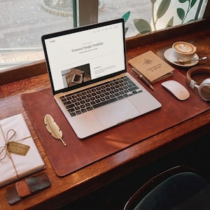 Peut inclure: Un sous-main en cuir marron foncé avec un ordinateur portable argenté, une souris sans fil blanche, un casque marron, un marque-page doré en forme de plume, un carnet à motif boussole, un stylo et un paquet blanc avec une étiquette "SABERBOOK VOL 1".
