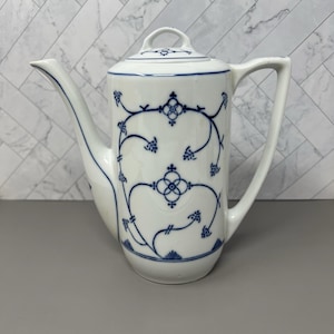 Peut inclure: Une cafetière en porcelaine blanche ornée d'un délicat motif de vigne florale bleue. La cafetière présente un corps cylindrique haut, un bec verseur incurvé, une anse robuste et un couvercle assorti avec un petit bouton. Le motif bleu est symétrique.