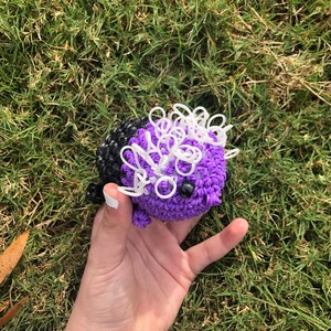 May include: A purple and black rubber band ball with white rubber bands on top. The ball is being held in a hand.