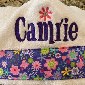 May include: White hooded towel with a purple embroidered name "Camrie" and a pink flower above the name. The towel has a purple and pink floral trim.