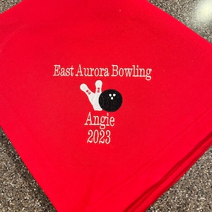 May include: Red fabric with white embroidery that says "East Aurora Bowling" with a bowling pin and ball graphic and "Angie 2023" underneath.