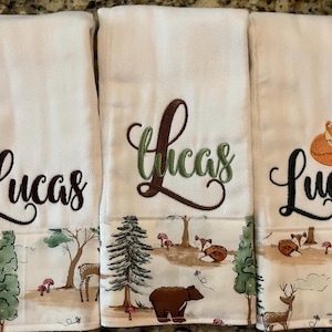 May include: Three white burp cloths with a woodland print border. Each cloth has the name "Lucas" embroidered in a different style. One cloth has the name in brown cursive, one in green cursive with a brown outline, and one in black cursive with a sleeping fox above the name.