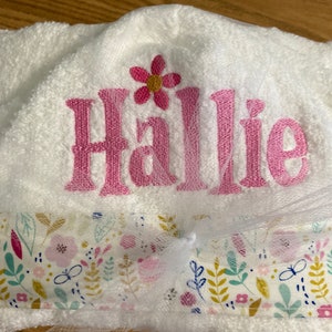May include: White hooded towel with a pink floral design and the name "Hallie" embroidered in pink. The towel has a floral print border with pink, yellow, and green flowers.