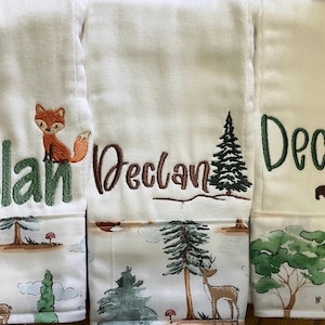 May include: White embroidered baby towels with the name "Declan" in green and brown thread. Each towel features a different woodland animal and landscape design, including a fox, bear, and deer.