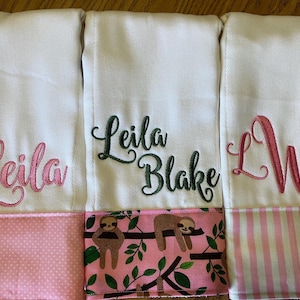 May include: Three white baby burp cloths with embroidered names and initials. One cloth has the name "Leila" in pink, another has "Leila Blake" in green, and the third has the initials "LWB" in pink. Each cloth has a patterned border.