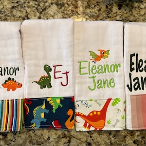May include: Four white burp cloths with dinosaur and bird embroidery. The cloths have different colored stripes and dinosaur patterns on the bottom.