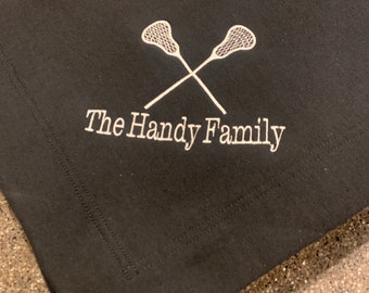 May include: Black fabric napkin with white embroidery of two lacrosse sticks crossed and the text "The Handy Family".