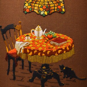 May include: Embroidered artwork depicting a table setting with a teapot, fruit, and a book. A chair sits beside the table, and two black cats are underneath. The scene is illuminated by a stained glass lampshade.