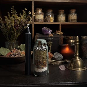 May include: A clear glass jar filled with layers of dried herbs, spices, and dark powder, sealed with sage green wax drips. A silver-toned pentacle charm with crescent moons hangs from the neck. Beside it stands a tall, smooth black candle.