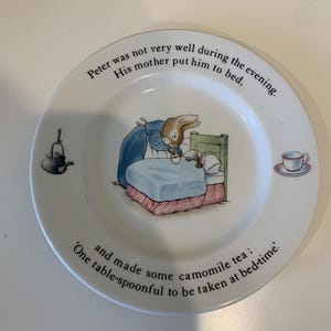 May include: A white ceramic plate featuring a Peter Rabbit illustration of a mother rabbit tucking a small rabbit into bed. The text reads: Peter was not very well during the evening. His mother put him to bed, and made some camomile tea; One table-spoonful to be taken at bed-time. A small kettle and a teacup are also illustrated.