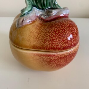 May include: A ceramic lidded container shaped like a pear with a glossy finish. The fruit features a gradient of yellow and red with a textured, speckled skin pattern. A sculpted green leaf sits atop the lid, adding a dimensional detail.