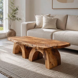 Op de afbeelding: Een lage salontafel gemaakt van een enkele plaat natuurlijk lichtbruin hout met een boomstamrand, ondersteund door twee dikke, gebogen houten poten. Het oppervlak vertoont een opvallende houtnerf en een gladde, gepolijste afwerking.