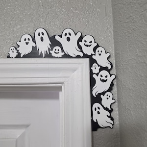 May include: A black L-shaped corner door frame decoration featuring a series of white ghost silhouettes with various facial expressions. The ghosts are arranged along the top and side edges of the black frame, creating a layered, spooky design.