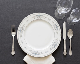 May include: A white porcelain dinner plate with a delicate blue and grey floral border pattern, accompanied by a stainless steel fork, knife, and spoon, plus two clear glass goblets and a folded white cloth napkin underneath the plate.