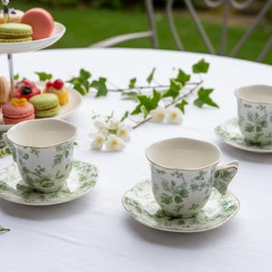 May include: Two white ceramic teacups and matching saucers decorated with a green botanical leaf pattern. The cups feature scalloped rims and handles, and are arranged on a white tablecloth alongside a two-tiered stand filled with colorful macarons.
