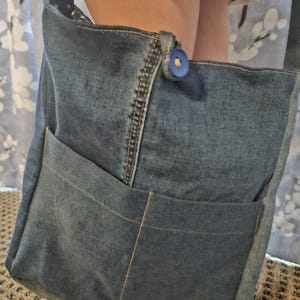 Op de afbeelding: Een denim schoudertas met een grote voorzak en een blauwe knoopsluiting. De tas is gemaakt van blauwe denim stof en heeft een zwarte band. De tas is ontworpen met zichtbare stiksels en een klassieke, casual stijl.