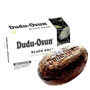 May include: A rectangular white box of Dudu-Osun black soap with black text and a palm leaf graphic, accompanied by an oval-shaped, dark brown, textured bar of soap with the brand name embossed on the surface. The box includes a scratch pin area.