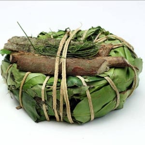May include: A round bundle of green leaves secured with light tan twine. Several brown wooden sticks are placed horizontally across the top of the leaf bundle, accompanied by a small cluster of thin, dark green pine-like needles.