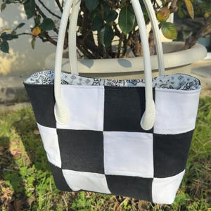 Peut inclure: Un sac fourre-tout à carreaux noirs et blancs avec des poignées blanches. L'extérieur du sac présente un motif de damier audacieux, tandis que la doublure intérieure présente un tissu à motifs. Les poignées sont blanches et semblent être en cuir.