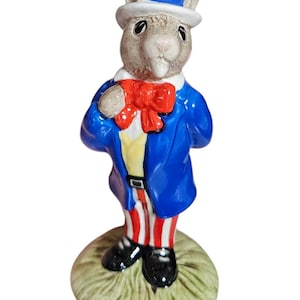 Può includere: Una statuetta in ceramica di un coniglio vestito da Zio Sam. Il coniglio indossa un cappotto blu, pantaloni a righe rosse e bianche e un papillon rosso. Il suo cappello è rosso, bianco e blu con una stella. La statuetta è su una base erbosa verde.