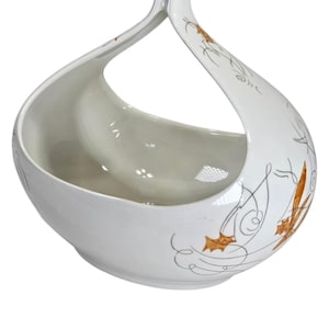 May include: White ceramic basket with a curved handle. The basket features a bowl shape with a decorative design of brown and gold leaf-like patterns and swirls. The handle curves up and over the bowl.
