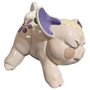 May include: A ceramic pig figurine in a playful pose, with closed eyes and rosy cheeks. The pig is primarily light pink, with purple accents on the ears and floral details. The figurine has a glossy finish and is designed to appear whimsical.