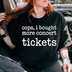 Könnte beinhalten: Ein schwarzes kurzärmeliges T-Shirt mit weißem Text auf der Vorderseite, der lautet: oops, i bought more concert tickets. Das T-Shirt hat einen Rundhalsausschnitt und eine Standardpassform, gefertigt aus einfarbigem schwarzem Stoff.