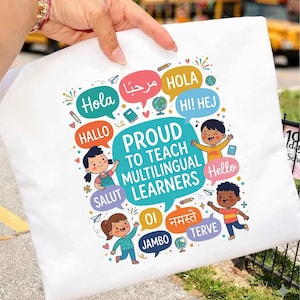 以下が含まれることがあります： PROUD TO TEACH MULTILINGUAL LEARNERSというテキストが書かれたカラフルなグラフィックが特徴の白いトートバッグ。デザインには、様々な言語の挨拶が入った吹き出し、子供たちのイラスト、地球儀、鉛筆が含まれています。