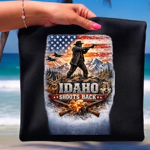 Peut inclure: Un t-shirt noir avec une impression graphique représentant une personne portant un chapeau et tenant une arme de poing, un drapeau américain, des avions de chasse, des montagnes, un cerf, un loup et deux fusils croisés. Le texte indique IDAHO SHOOTS BACK.